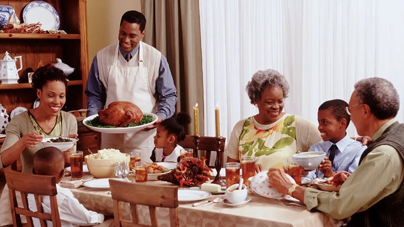 A joyful multi-generational African American family gathered around a dining table, sharing a Thanksgiving meal with a roasted turkey as the centerpiece.