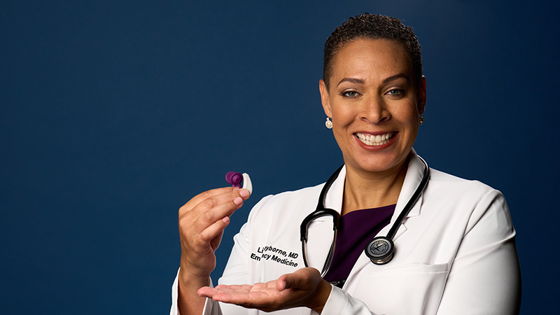 A smiling Black female doctor, Dr. L. Lilyborne, wearing a white lab coat and holding a small medical device resembling an inhaler, with a dark blue background.