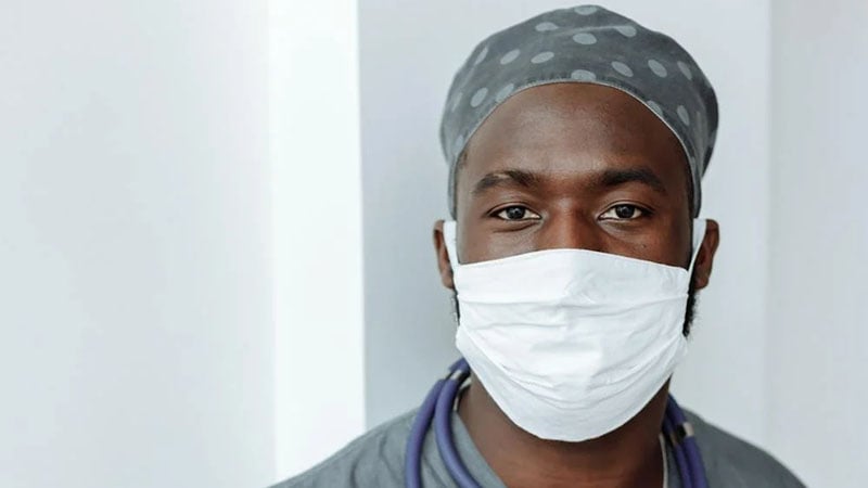 A thoughtful-looking Black male doctor in a white coat with a stethoscope around his neck stands with his arms crossed in a hospital hallway.
