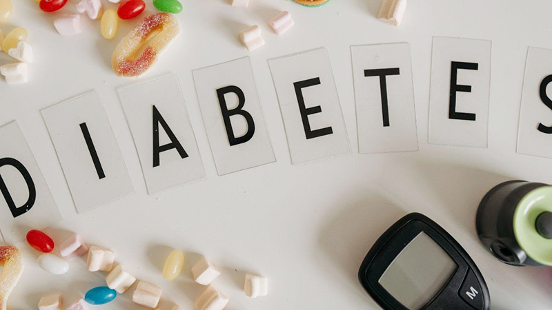 Word 'DIABETES' spelled out with letter cards on a white surface, surrounded by colorful candies, marshmallows, a glucose meter, and a lancing device.