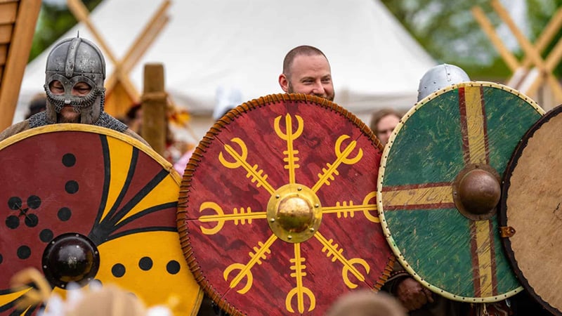 A vibrant scene of Viking Fest featuring participants in historical costumes with round shields, set against a backdrop of tents and wooden structures.