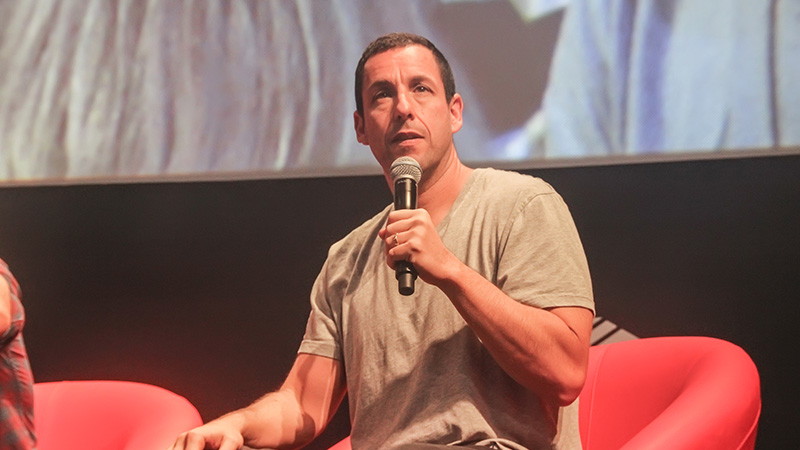 Adam Sandler speaking into a microphone during a public event, seated on a red chair with a casual grey t-shirt.