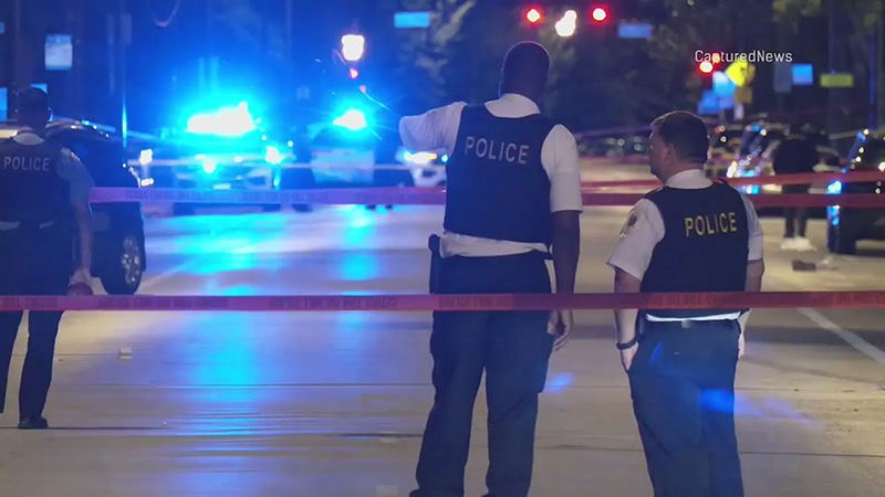 Police officers stand behind crime scene tape at night, illuminated by flashing blue and red lights, symbolizing public safety challenges in Chicago.