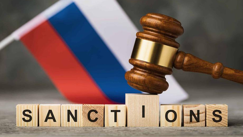 Wooden blocks spelling "SANCTIONS" with a gavel resting on them, set against a blurred Russian flag background.