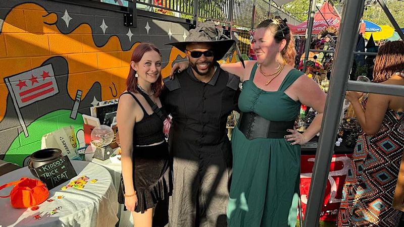 Three people in costume at an outdoor Halloween-themed event, standing in front of a mural and vendor stalls.