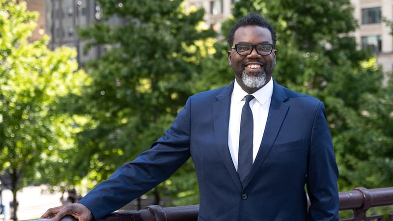 A smiling man in a dark blue suit and glasses standing outdoors with greenery and urban buildings in the background.
