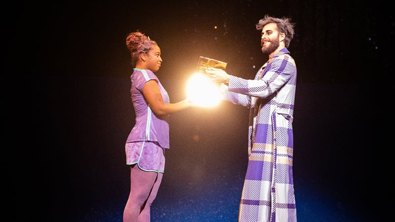 Two performers on stage holding a glowing object, surrounded by dramatic lighting, representing a scene from Cirque du Soleil's holiday show.
