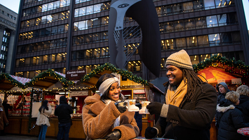 A young couple toasting with festive mugs at the Christkindlmarket in Chicago, surrounded by decorated wooden stalls and holiday lights.