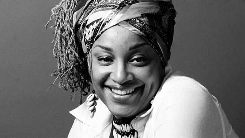 A black-and-white portrait of Jade Harrell, smiling warmly, wearing a patterned headwrap, shell jewelry, and a white collared shirt.