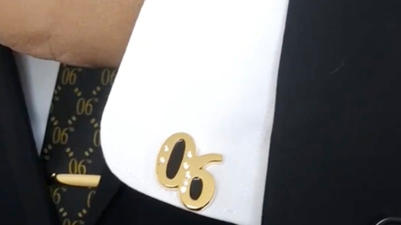 A close-up of a white shirt cuff adorned with a gold Alpha Phi Alpha cufflink, paired with a black suit and tie featuring gold accents.