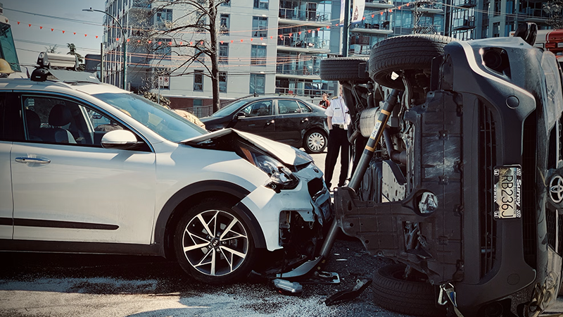 A car accident scene showing a white SUV with front-end damage and an overturned dark gray SUV on a city street.