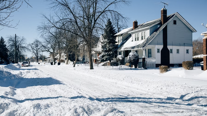 Snow-covered suburban street in Detroit during winter, with houses, bare trees, and evergreens lining the partially plowed road under a clear blue sky."