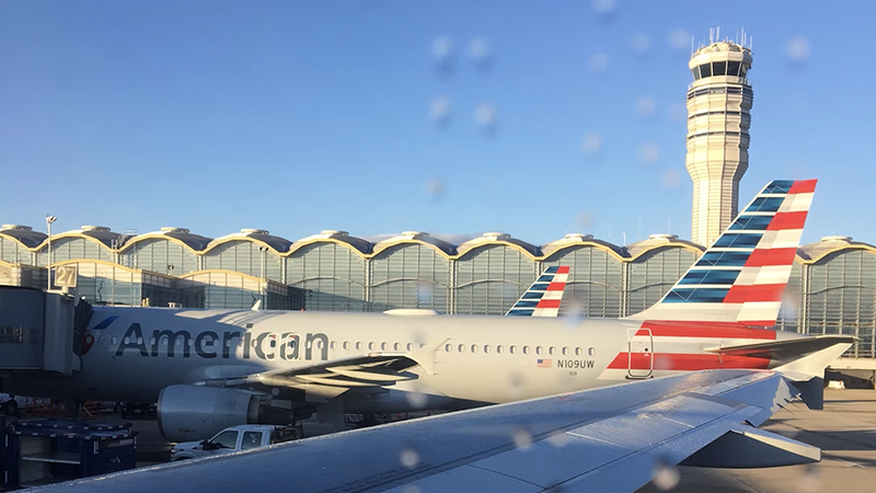 An American Airlines plane at a DC-area airport gate, with a modern terminal and control tower in the background under a clear blue sky.