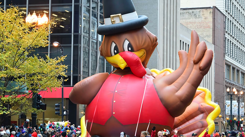 A giant inflatable turkey balloon wearing a top hat and red vest floats above a crowd during a festive Thanksgiving parade in Chicago.