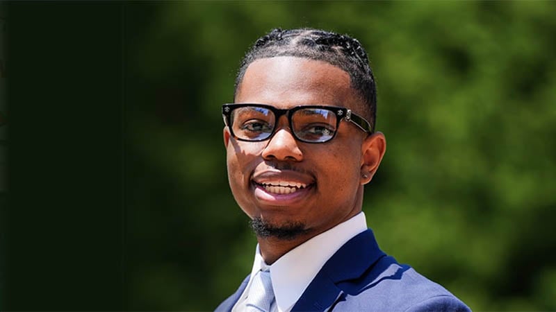A young Black entrepreneur in a suit and tie, smiling confidently against a blurred green background.