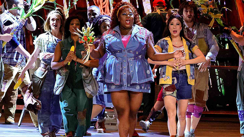 Performers on stage in vibrant costumes during a musical, featuring a rustic theme with corn stalks and expressive poses.