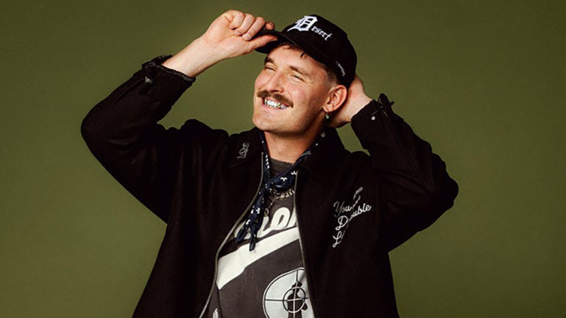 A man smiling broadly, wearing a black 'Desert' cap, dark jacket, and a bandana, with a Public Enemy logo on his T-shirt, set against an olive-green background.