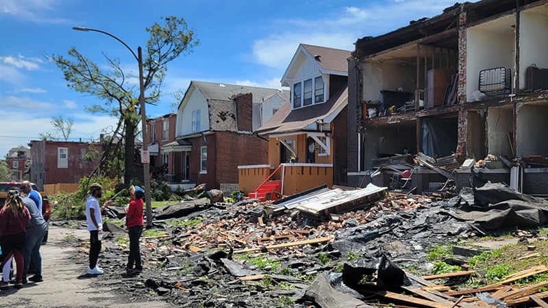A tornado-damaged neighborhood in St. Louis with debris, damaged homes, and residents surveying the aftermath under a clear blue sky