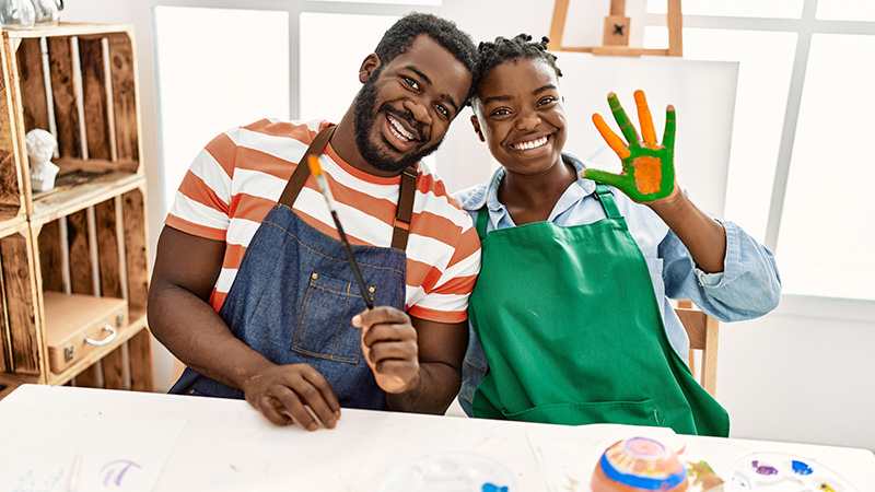 A smiling man and woman in an art studio, engaged in painting. The man holds a paintbrush, and the woman shows her hand painted with green and orange.