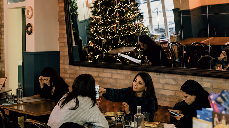 A cozy restaurant interior during the holiday season, featuring patrons at tables, a large mirror reflecting a decorated Christmas tree, and warm lighting.