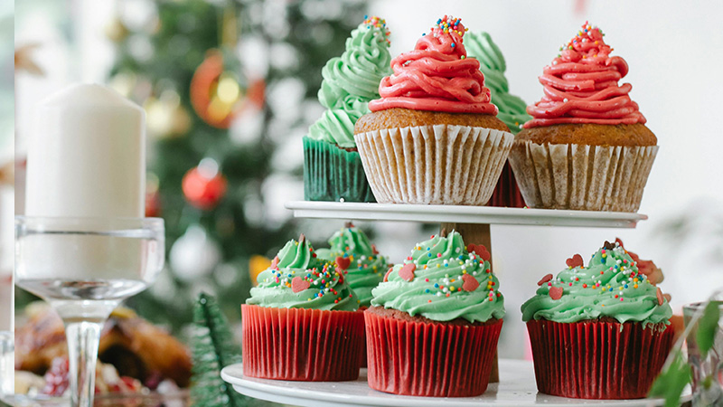 Festive gingerbread cupcakes with red and green frosting, sprinkles, and heart-shaped candies on a tiered stand with a Christmas tree in the background.