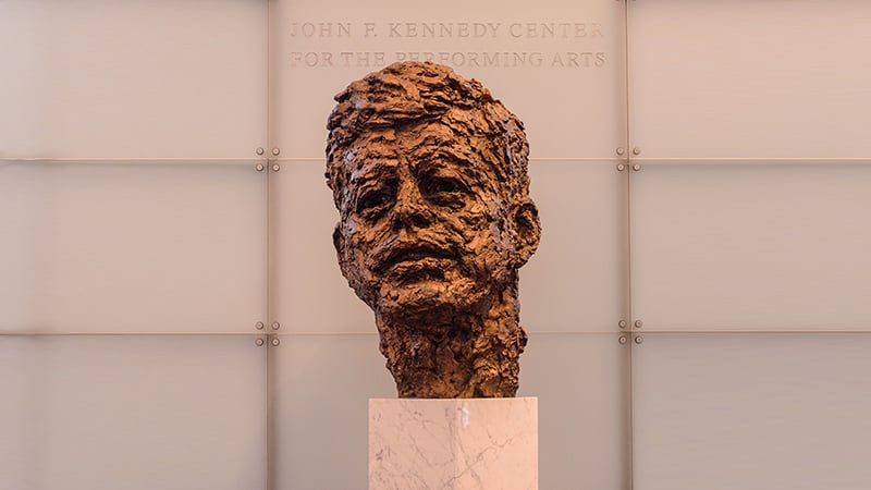 Bronze bust of John F. Kennedy on a marble pedestal at the Kennedy Center, with a frosted glass wall and engraved text in the background.