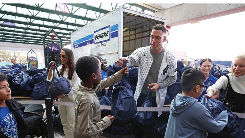 New York Yankees player Aaron Judge handing a blue gift bag to a young boy near a Penske truck during an MLB Cares holiday community event.
