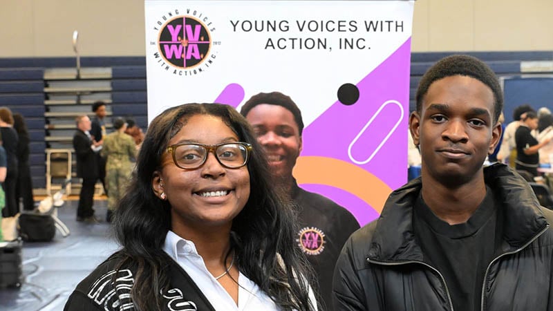 Two young individuals standing in front of a banner for Young Voices with Action, promoting youth empowerment and leadership.