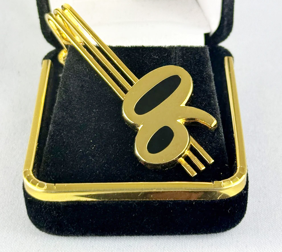 The Alpha 06 tie bar displayed in a black velvet box, highlighting its golden finish and symbolic design.