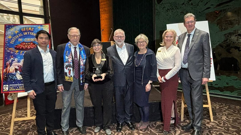 Jessica Hentoff, holding a translucent award plaque, stands with six other formally dressed individuals on a stage during the 48th International Circus Festival in Monte Carlo. A colorful circus-themed poster is visible in the background.