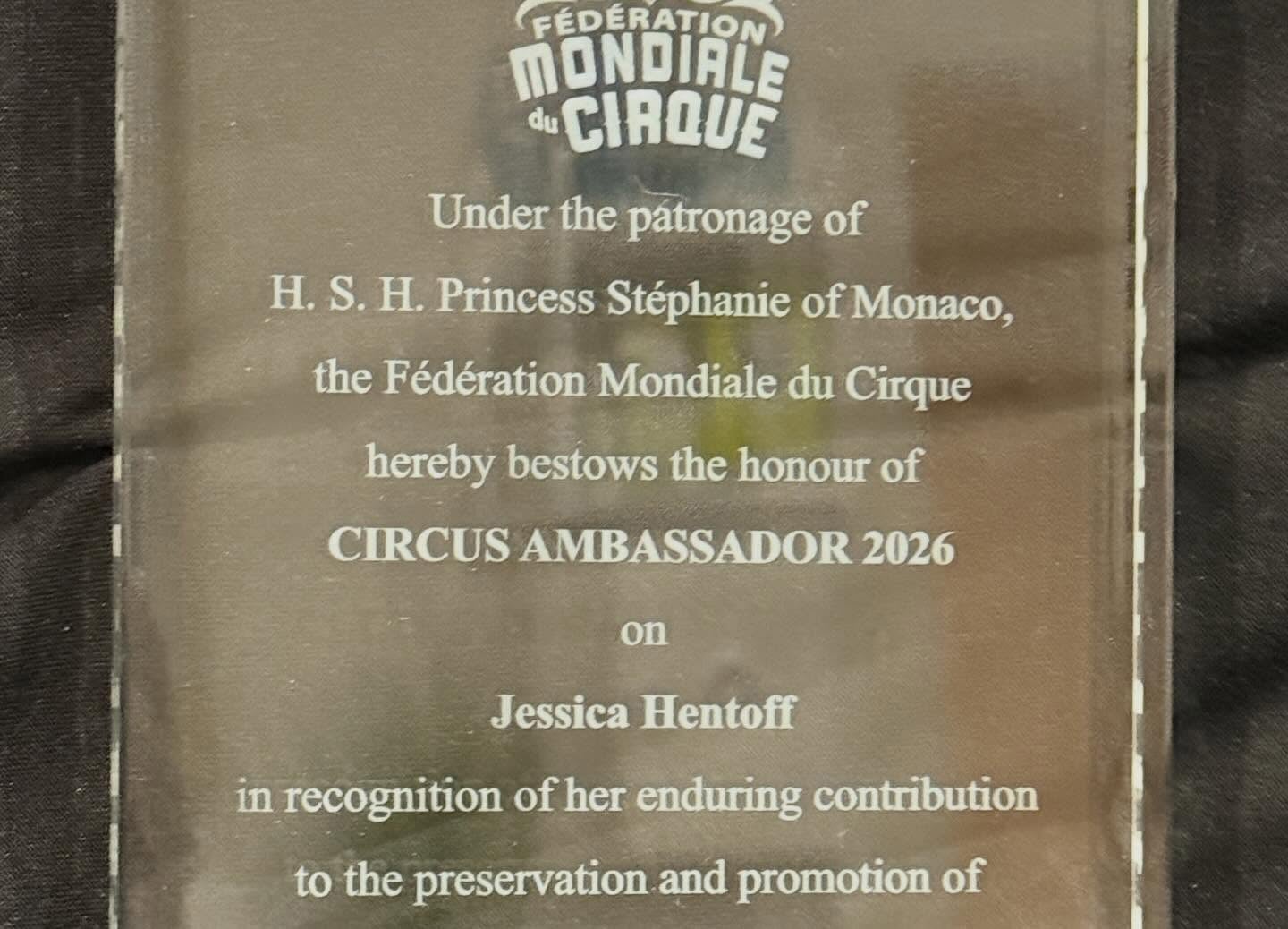 A clear rectangular award plaque with engraved text from the Fédération Mondiale du Cirque, recognizing Jessica Hentoff as Circus Ambassador 2026 under the patronage of H.S.H. Princess Stéphanie of Monaco. The plaque is set against a dark, textured background.