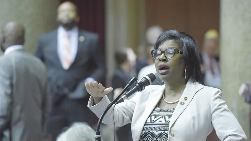 Missouri Senator Karla May speaking passionately at a legislative session, wearing a light blazer and glasses, with a microphone in front of her.