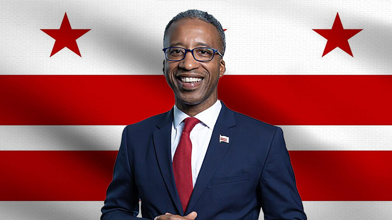 Kenyan McDuffie smiling in a suit with the Washington D.C. flag in the background.