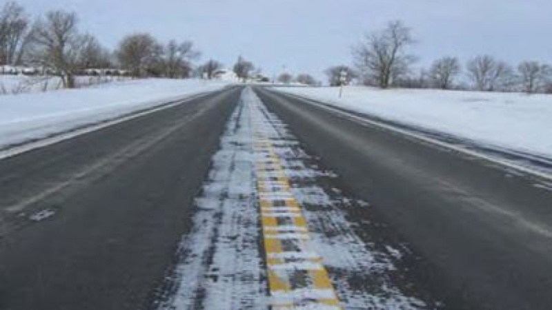Snow-covered Missouri road with a double yellow line, surrounded by bare trees under an overcast sky.