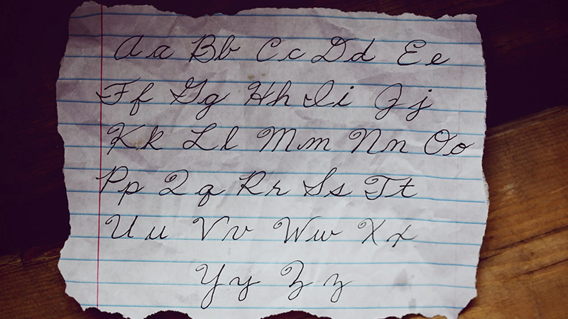 Torn lined paper with cursive alphabet handwritten in black ink, placed on a wooden surface with warm lighting.