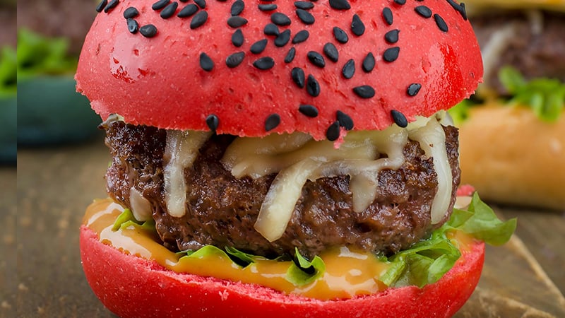 Bright red-bun mini-burger with beef patty, melted cheese, lettuce, and sauce on a rustic wooden surface.