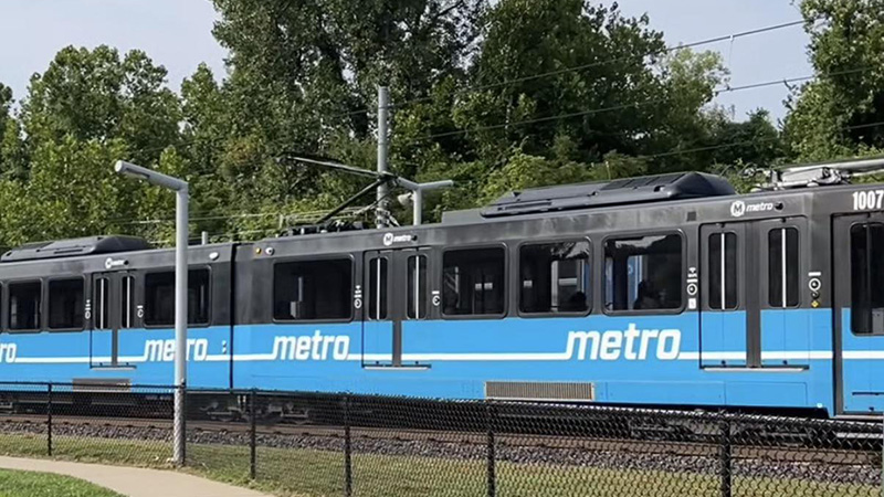 A blue and gray metro train with the number "1007" on its side, running on tracks next to a chain-link fence and trees.