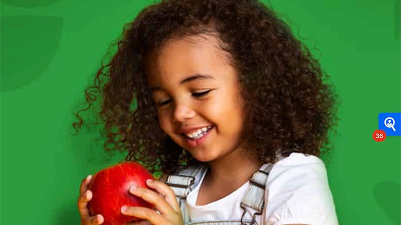A young girl with curly hair smiles while holding a red apple against a vibrant green background, symbolizing health and joy.