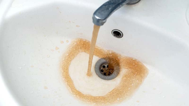 A stream of dirty, brown water flows from a silver faucet into a white ceramic sink, indicating a water contamination problem.