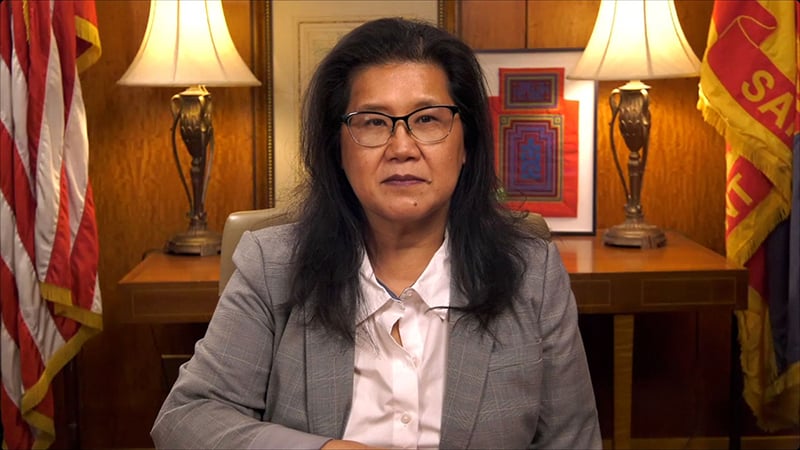 A professional portrait of an Asian woman wearing glasses and a blazer, seated in an office setting with wooden paneling, flags, and table lamps in the background.
