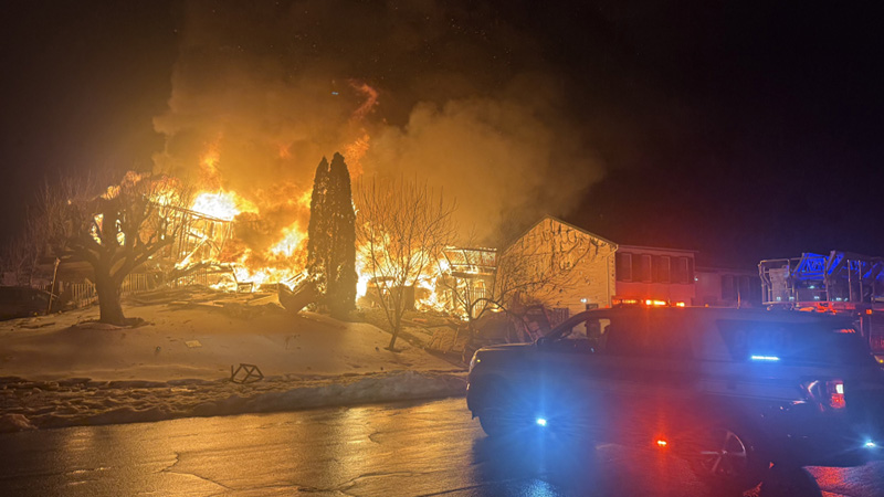 A house at night is completely engulfed in bright orange and yellow flames, with an emergency vehicle visible in the foreground.