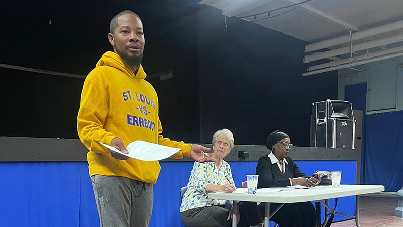 The image shows Alderman Rasheen Aldridge addressing a community meeting. He is wearing a yellow hoodie and gesturing while holding papers, with two women seated at a table in the background.