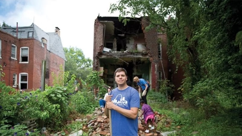 A man wearing a blue t-shirt with "I LOVE ST. LOUIS" printed on it stands amidst rubble and overgrown vegetation, holding a tool. In the background, dilapidated brick buildings and other individuals, including a child, are engaged in cleanup activities