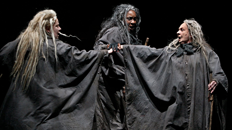 Three actors portraying witches in Macbeth at the Guthrie Theater, dressed in dark cloaks with dramatic lighting.