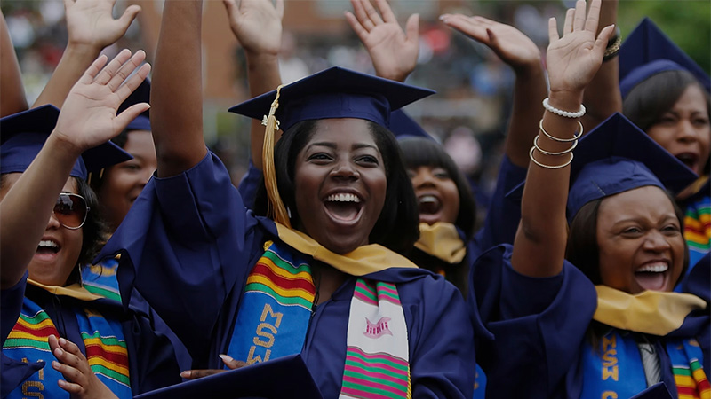 A group of young Black women in navy blue graduation gowns and caps with gold tassels celebrate joyfully at a graduation ceremony. The central figure wears a gold stole and a colorful kente cloth-like stole with "MSM C" visible on it. Arms are raised in celebration, and the atmosphere is vibrant and jubilant.