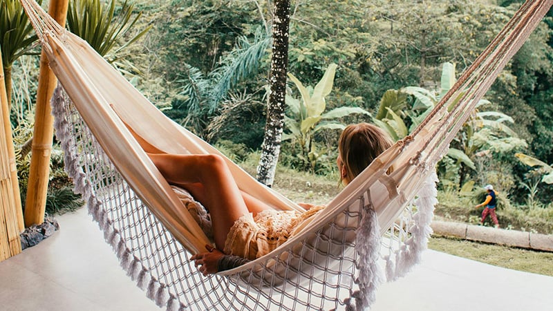 A woman relaxes in a cream-colored hammock with tassels, suspended between bamboo poles on a light patio. She gazes at a lush green tropical landscape with dense foliage and a distant figure walking on a path.