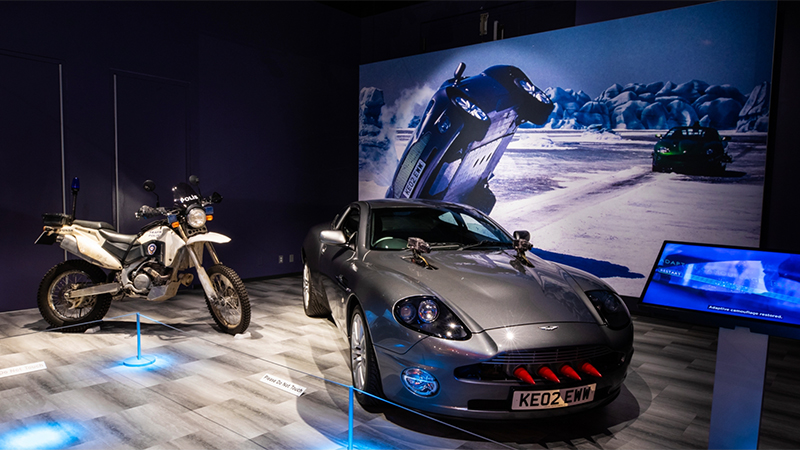 A museum exhibit featuring a white police motorcycle labeled "POLIS" and a gray Aston Martin Vanquish with red weapon barrels in its grille. The backdrop shows an icy landscape with a car flipping mid-air, referencing a James Bond movie scene.