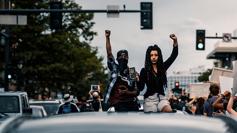 Two individuals stand on a vehicle with raised fists during a protest, surrounded by a crowd and urban street elements, including traffic lights and a "Black Lives Matter" sign.