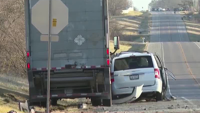 A roadside accident involving a white minivan and a gray semi-trailer on a rural road, with visible damage to the minivan's front-right side.