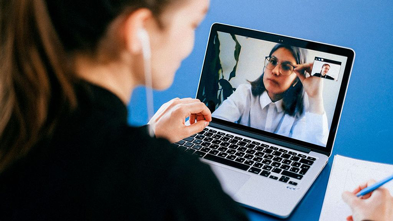 A person participating in a video call on a laptop, taking notes on a blue desk, symbolizing remote work and virtual meetings.